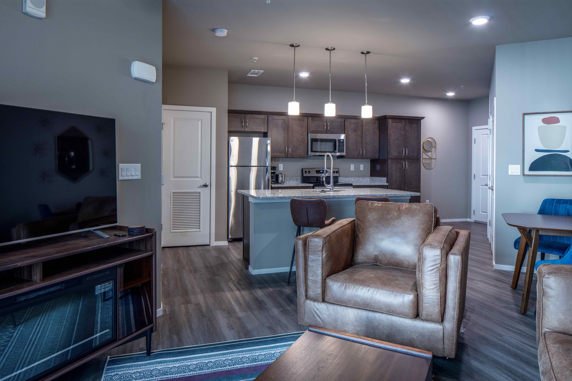 Modern living room with a kitchen, brown leather armchairs, and a dark wood cabinet.