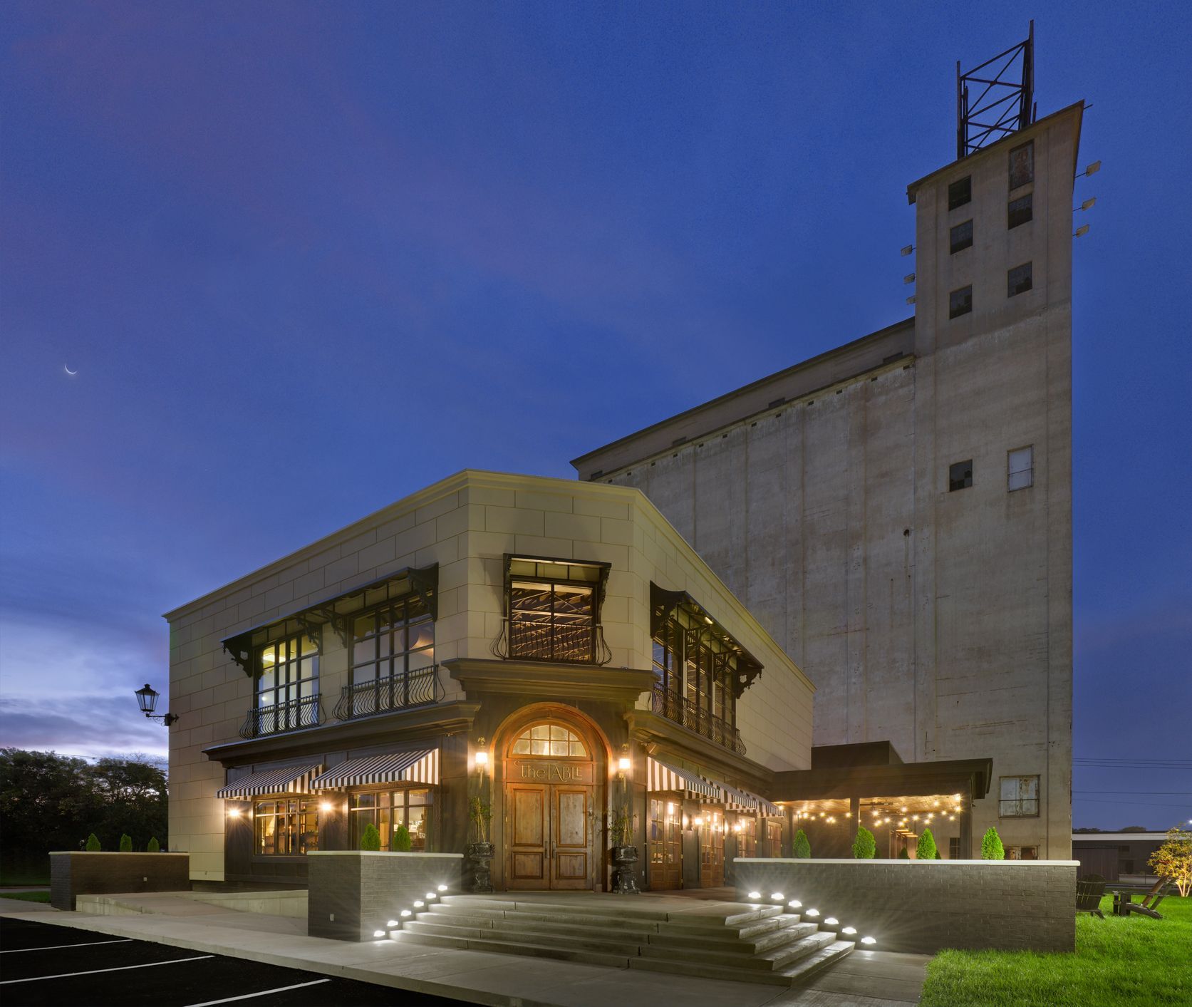 A modern restaurant building with warm outdoor lighting, attached to a tall, industrial concrete grain silo at dusk.
