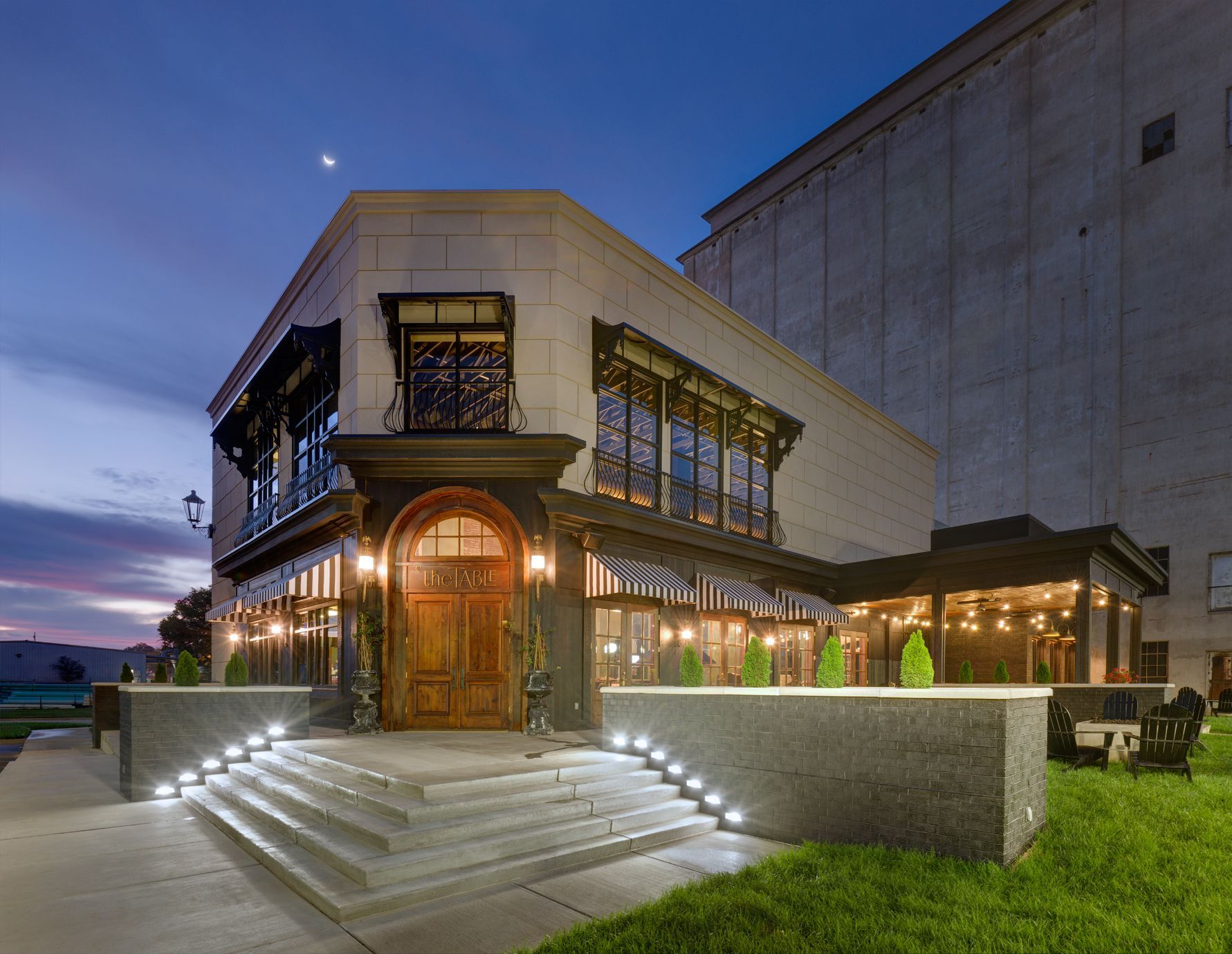A dimly lit, ornate building exterior at dusk, featuring stone steps with accent lighting leading to a wooden arched door.