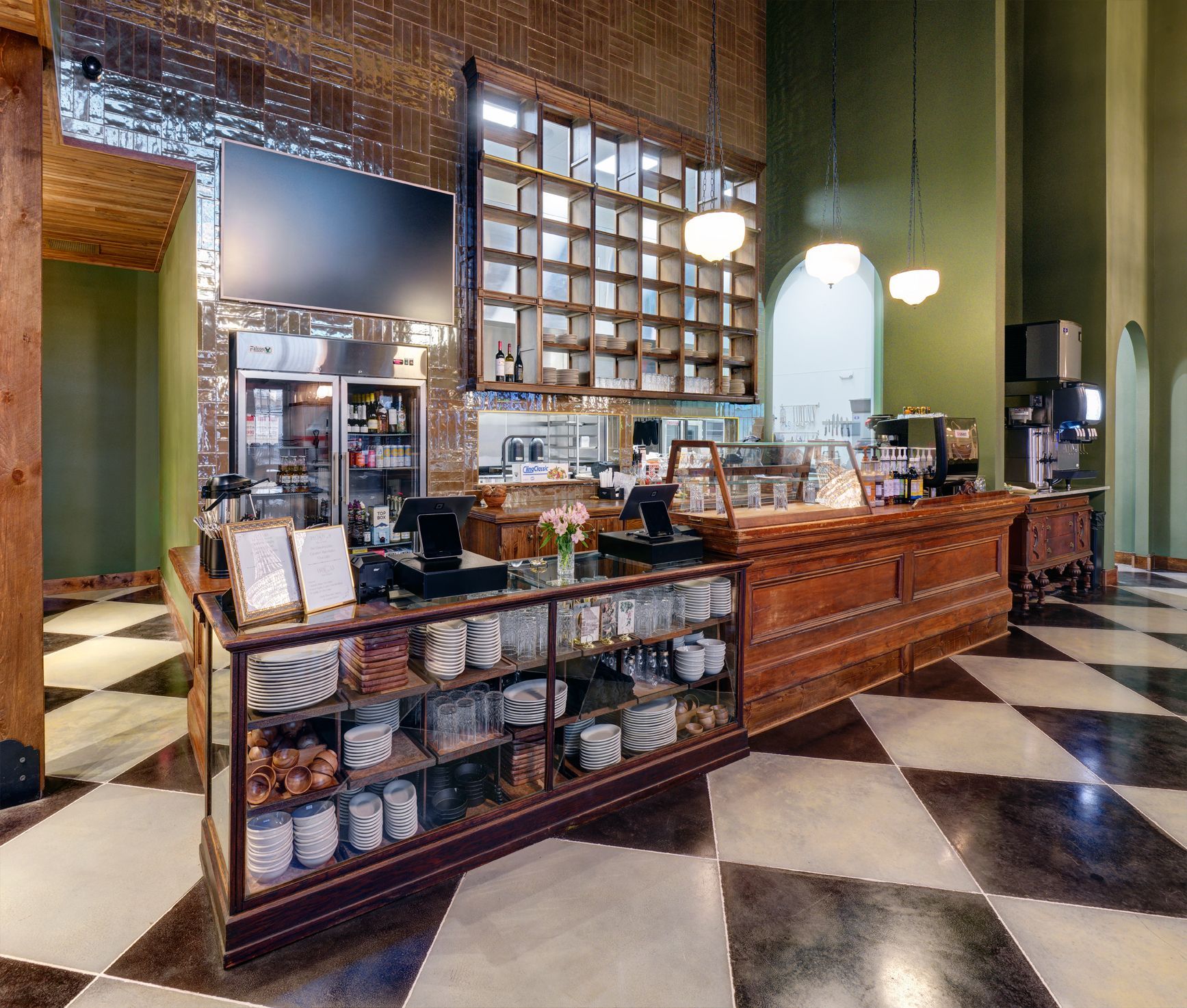 A bakery or coffee shop counter with wooden shelving, a glass display case of goods, and a checkered floor.