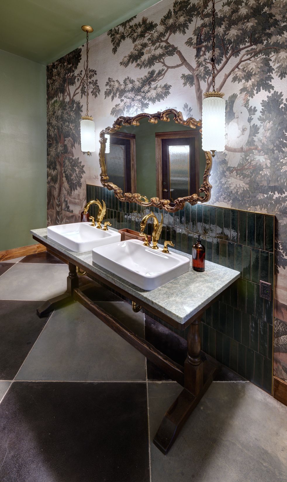 Double vanity with white vessel sinks and gold faucets against a scenic wallpaper wall with an ornate gold mirror.