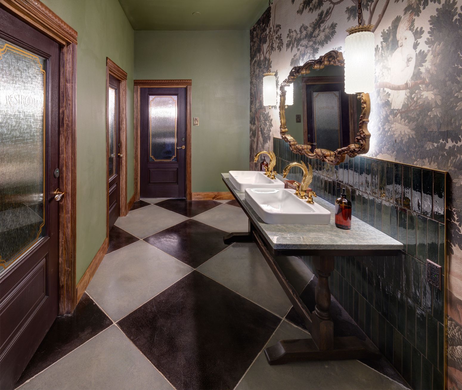 A hotel restroom with a checkered floor, dual sinks on a wooden vanity, and patterned wallpaper behind an ornate mirror.