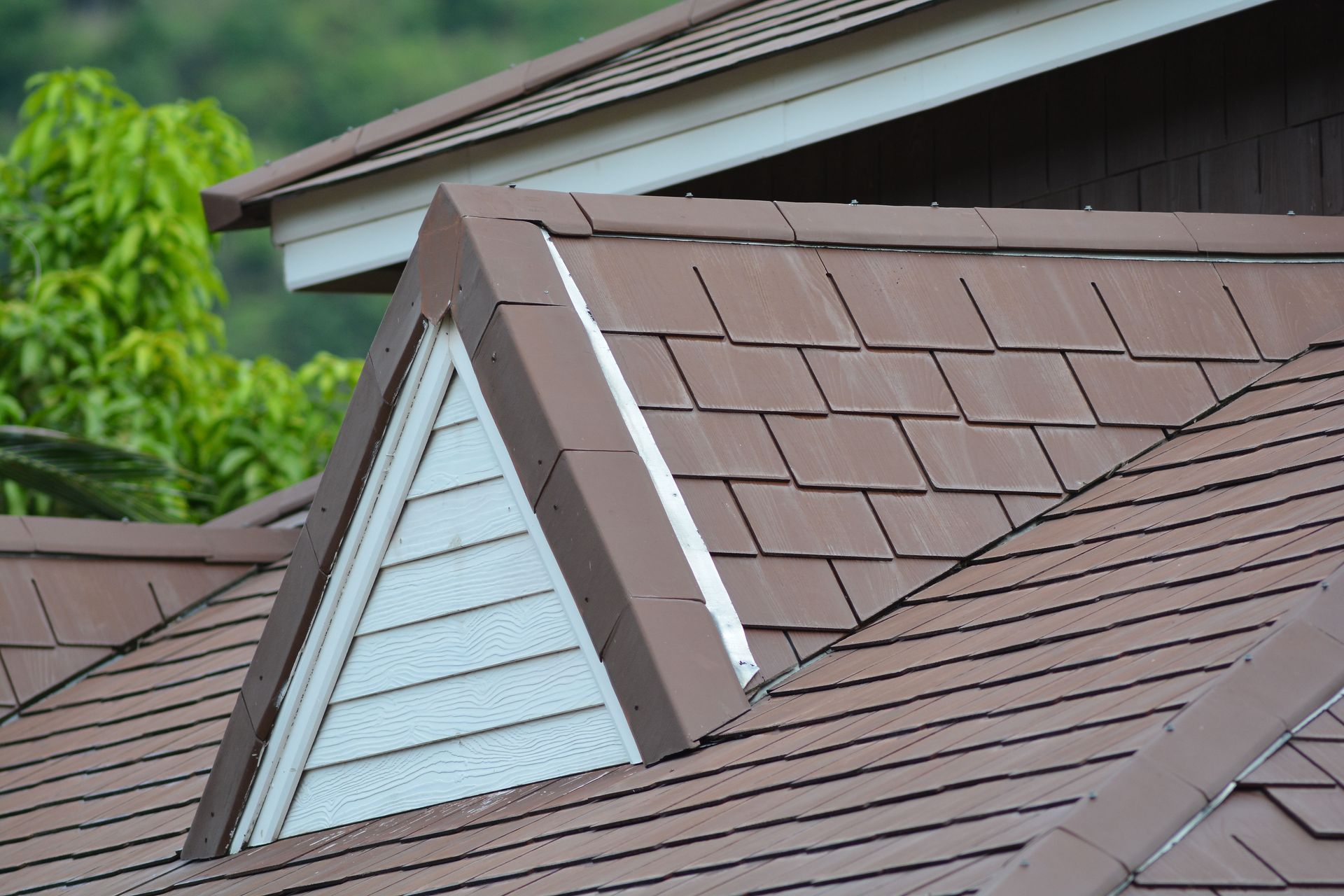 A triangular dormer window set into a brown tiled roof with white siding, under a green-foliaged sky.