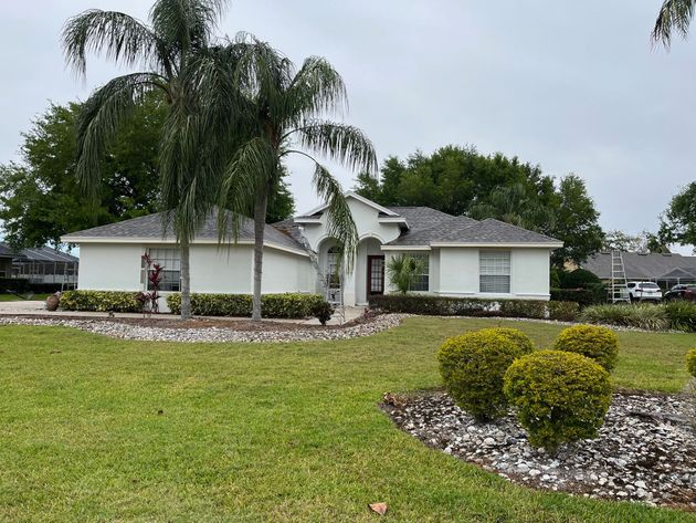 A white, single-story house with a grey shingled roof, palm trees, and landscaped bushes under a cloudy sky.