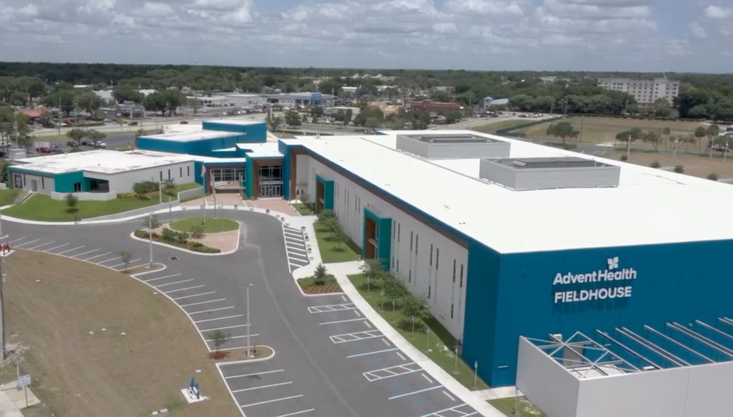 Aerial view of the AdventHealth Fieldhouse with a white roof and teal walls, situated next to a parking lot and grassy area.