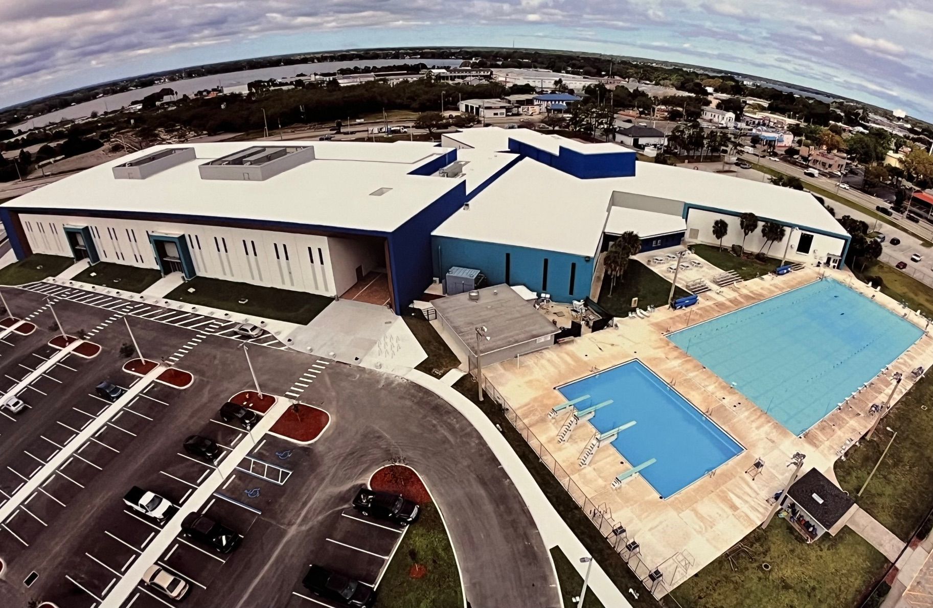 Aerial view of a large white and blue facility with a parking lot and two outdoor swimming pools.