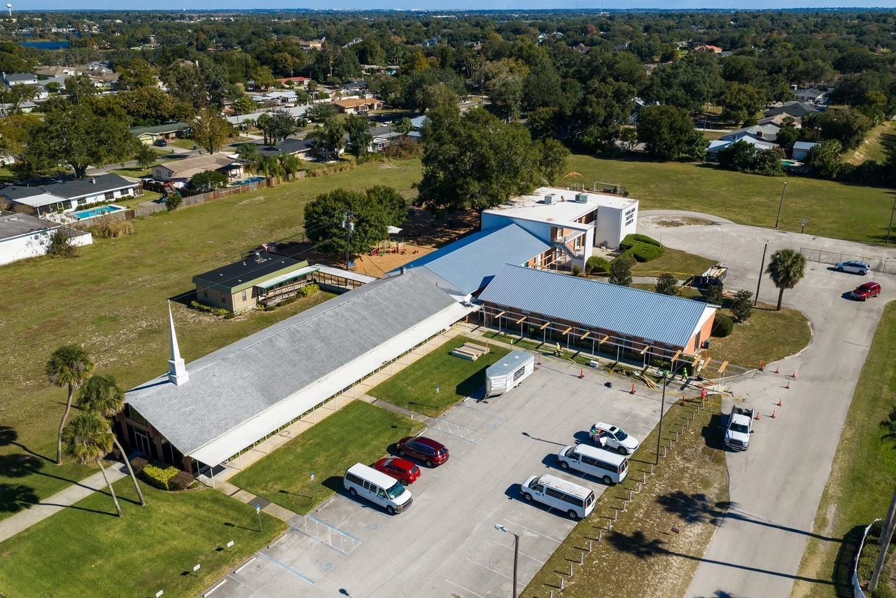 Aerial view of a church complex with grey roofs, surrounding parking areas, and trees, situated in a suburban neighborhood.