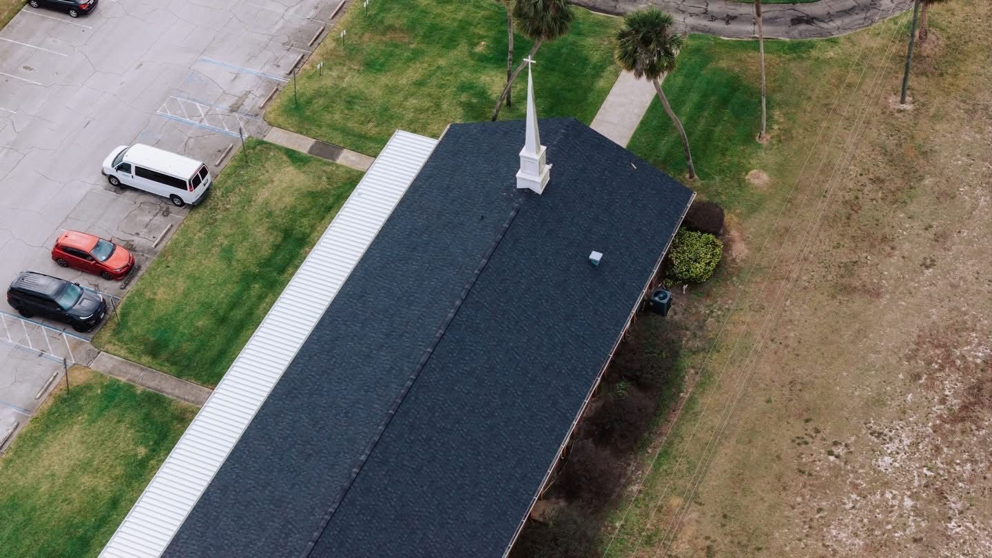 An aerial view shows a dark-roofed building with a white steeple next to a parking lot with several vehicles.