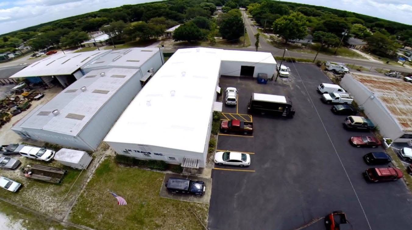 Aerial view of an L-shaped commercial building and parking lot with several vehicles surrounded by trees.