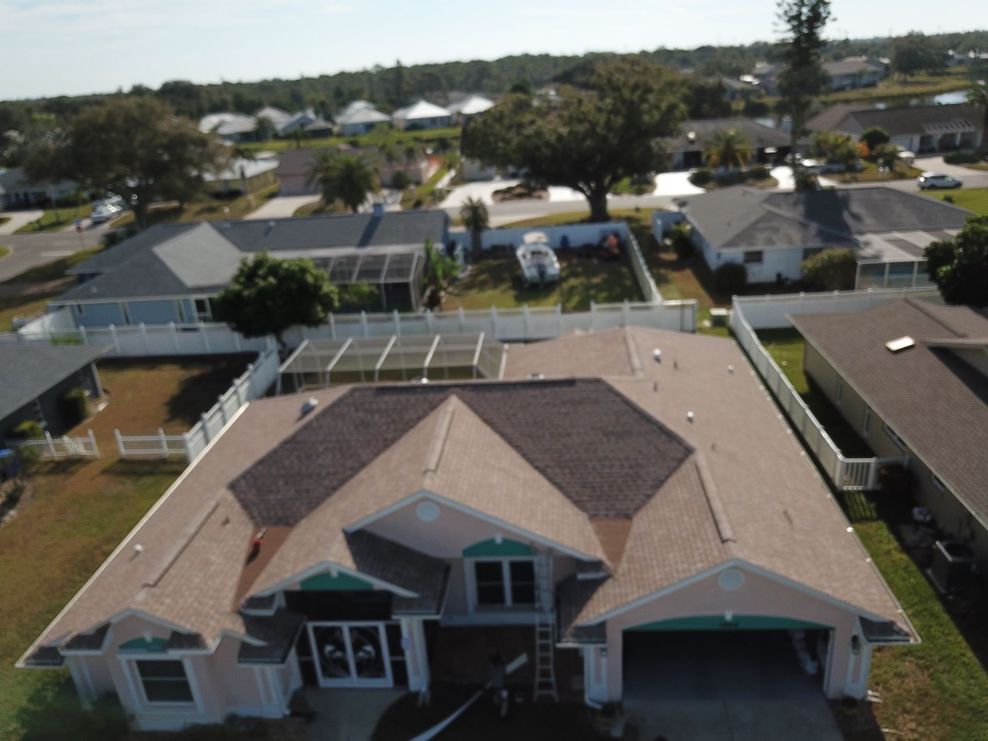 Aerial view of suburban homes with brown roofs, white fences, and green lawns on a sunny day.