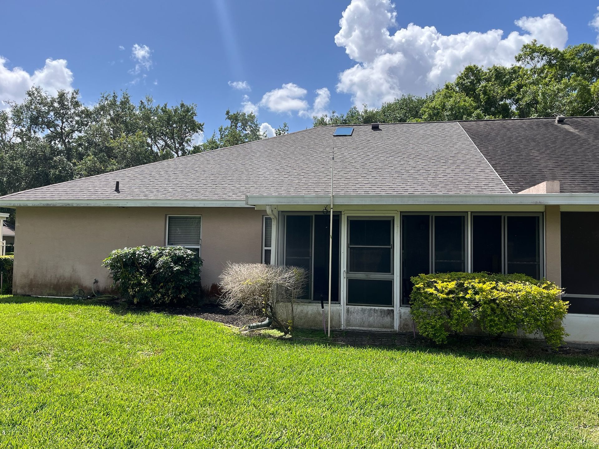 Tan house with a dark, algae-covered roof, screened-in porch, and green lawn under a partly cloudy sky.