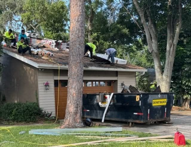 Roofers replacing shingles on a garage roof. Dumpster and debris near the garage, with a large tree in the foreground.