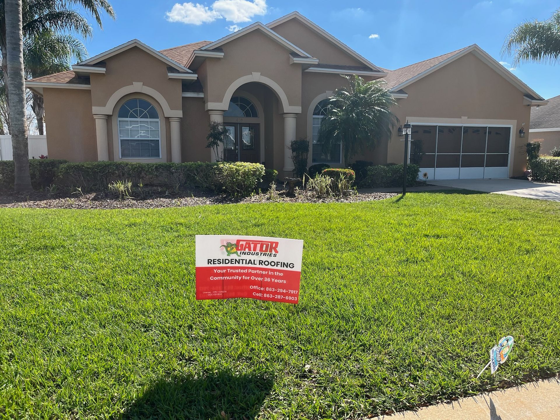 House with sign in the yard. Beige stucco, green lawn, palm trees, and a sign for roofing services.