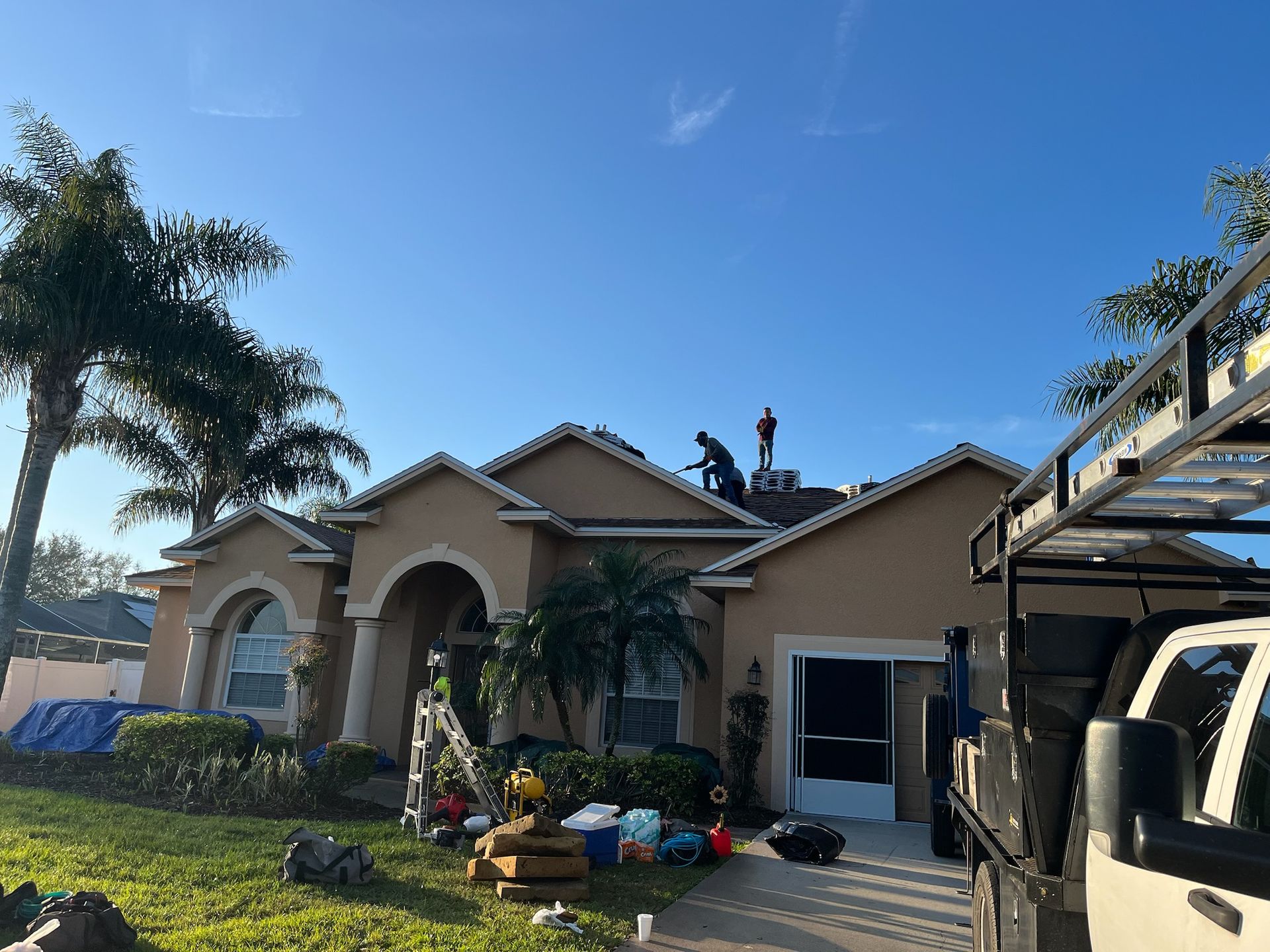 Workers on a roof, replacing shingles on a house with a tan exterior under a clear blue sky.