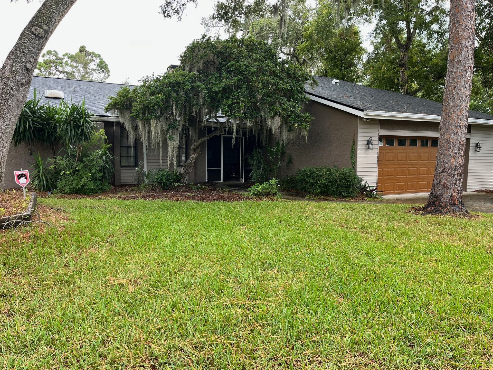 Ranch-style house with tan stucco, green lawn, brown garage door, and trees with Spanish moss.