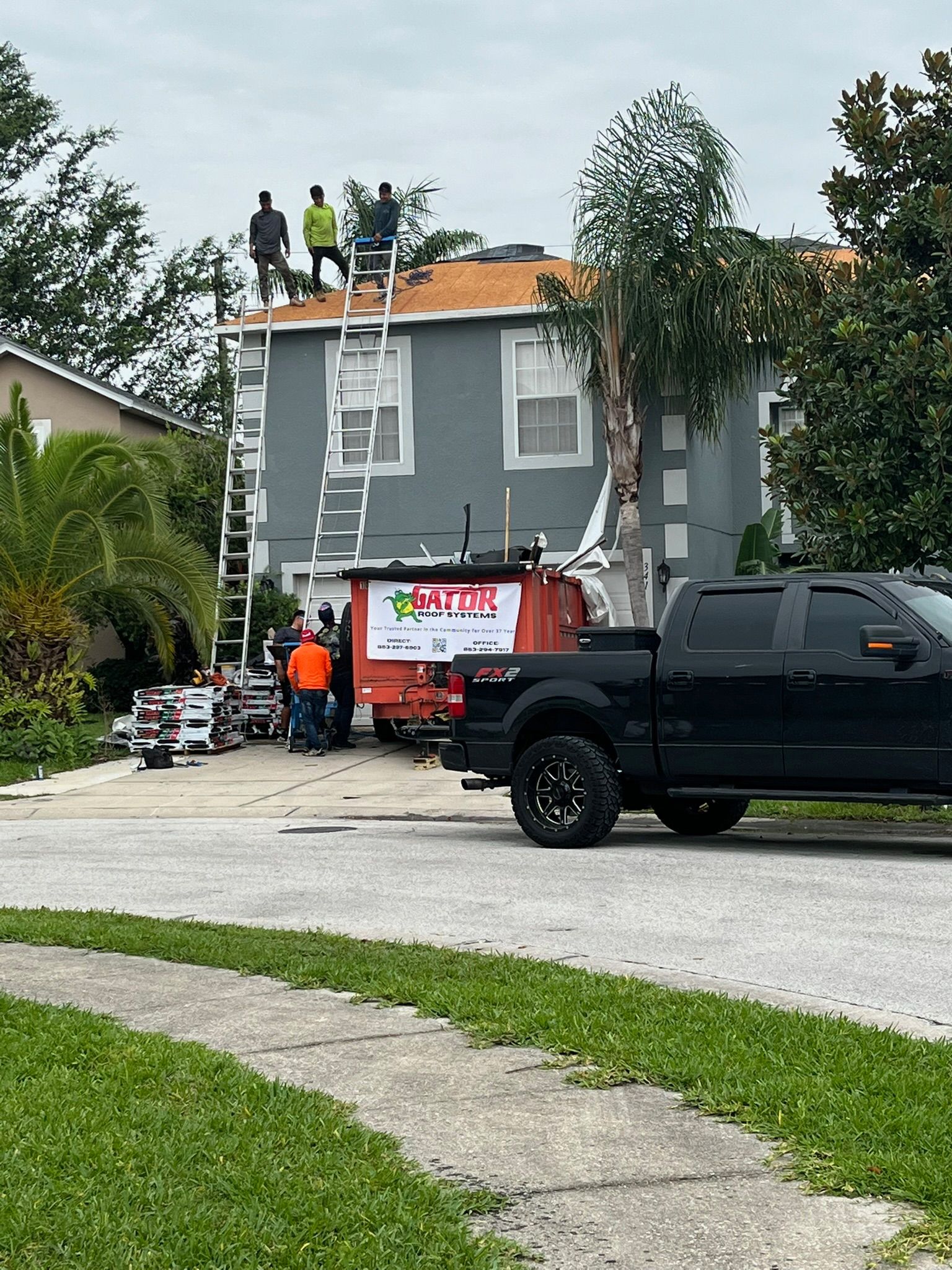 Roofers on a house with ladders and equipment, orange truck, black pickup, cloudy sky.