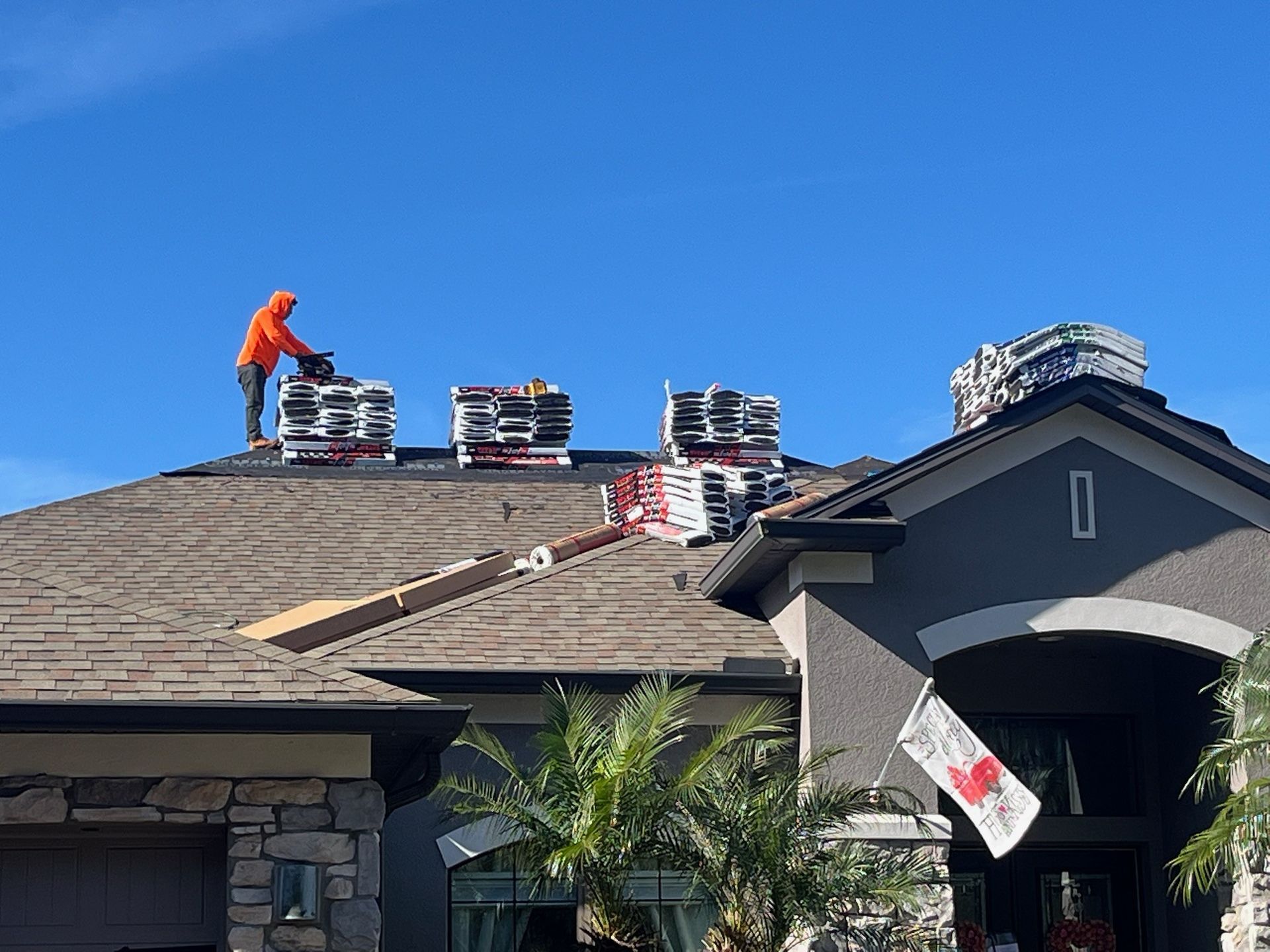 Roofer in orange jacket works on a roof, surrounded by bundles of shingles, under a blue sky.