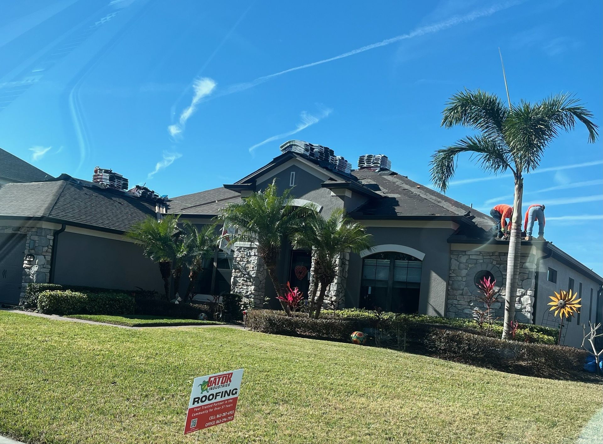 House with dark roof, gray stone facade, palm tree, and sign on lawn under a blue sky.
