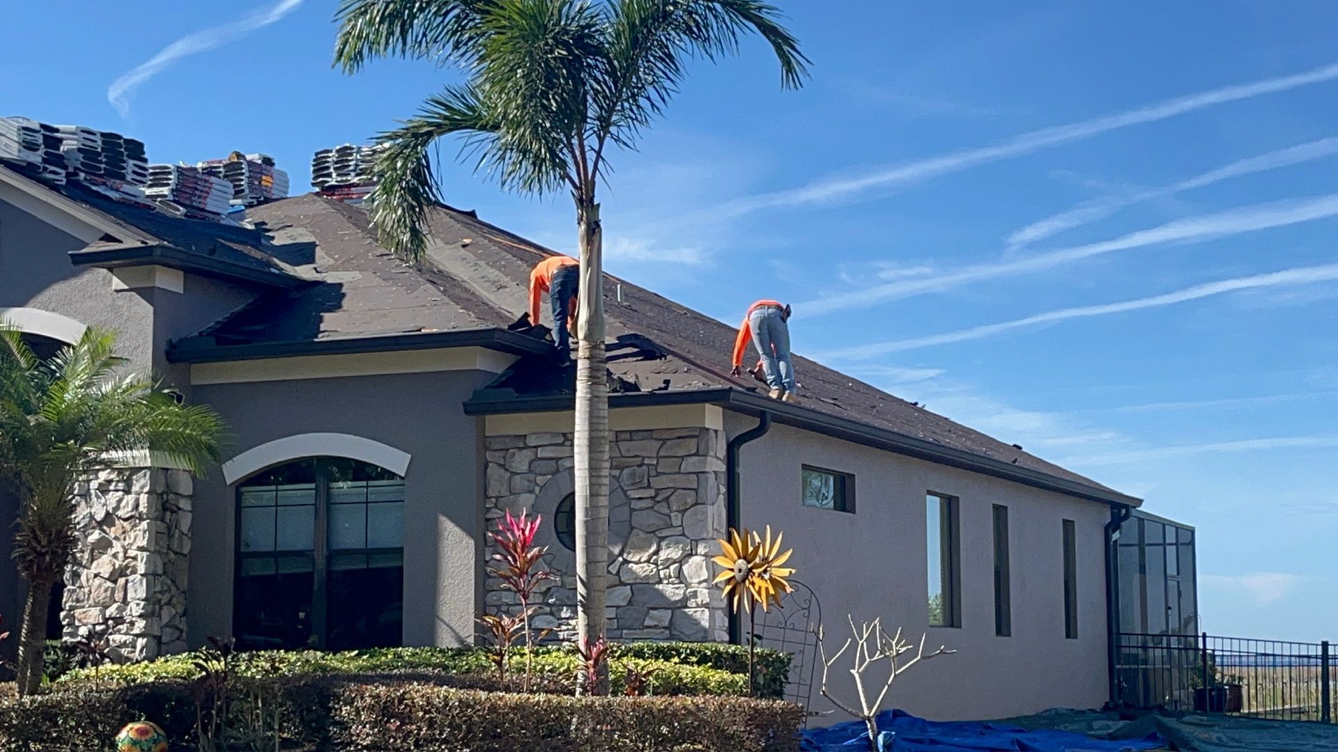 Two roofers on a residential roof, working. Blue sky. Palm tree and stone accents.