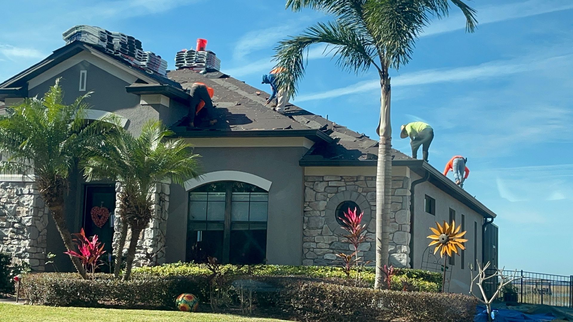Roofers on a gray house with a stone facade, palm trees, and decorative flowers.