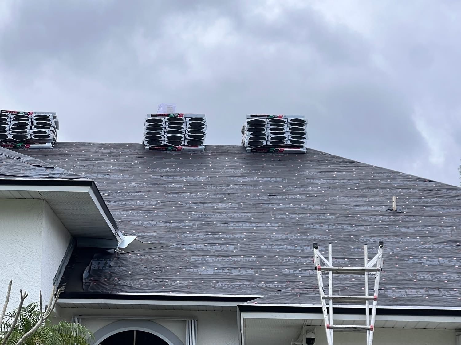 Roof with bundles of shingles, ladder, overcast sky.