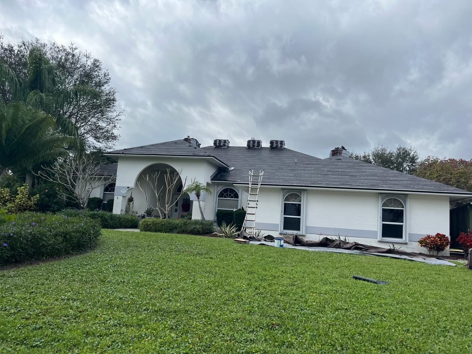 House with dark gray roof being worked on, ladder against roof, cloudy sky.