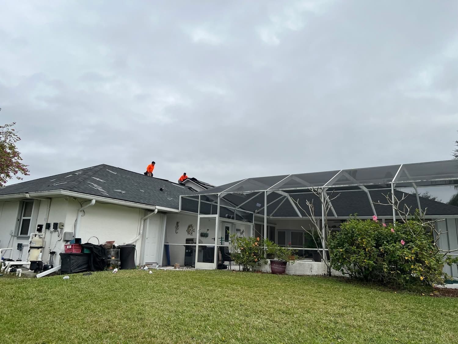 A person on a roof in an orange vest, cloudy sky, single-story house, screened-in porch, green grass.