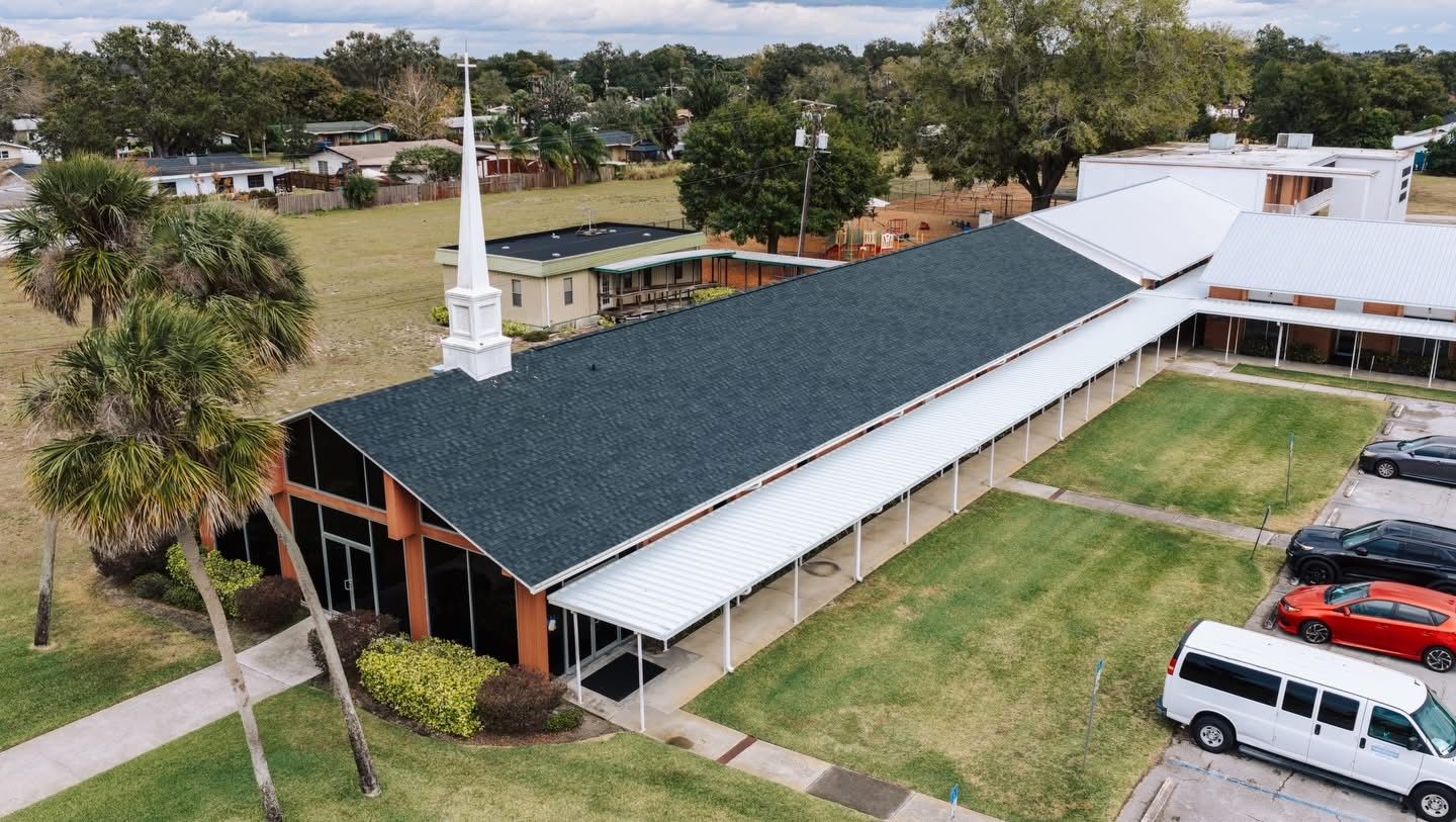 Church building with a white steeple and long, dark roof; cars parked nearby.