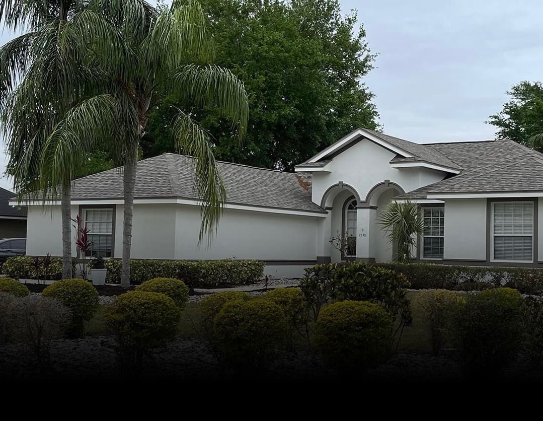 White house with gray roof, shrubs in front, palm trees on the left.