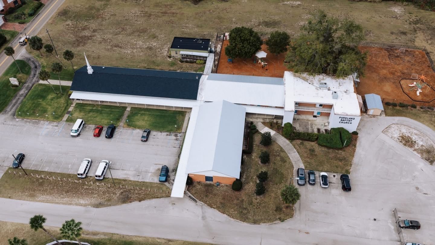 Aerial view of a church with a long, multi-sectioned building, parking lot, and surrounding greenery.