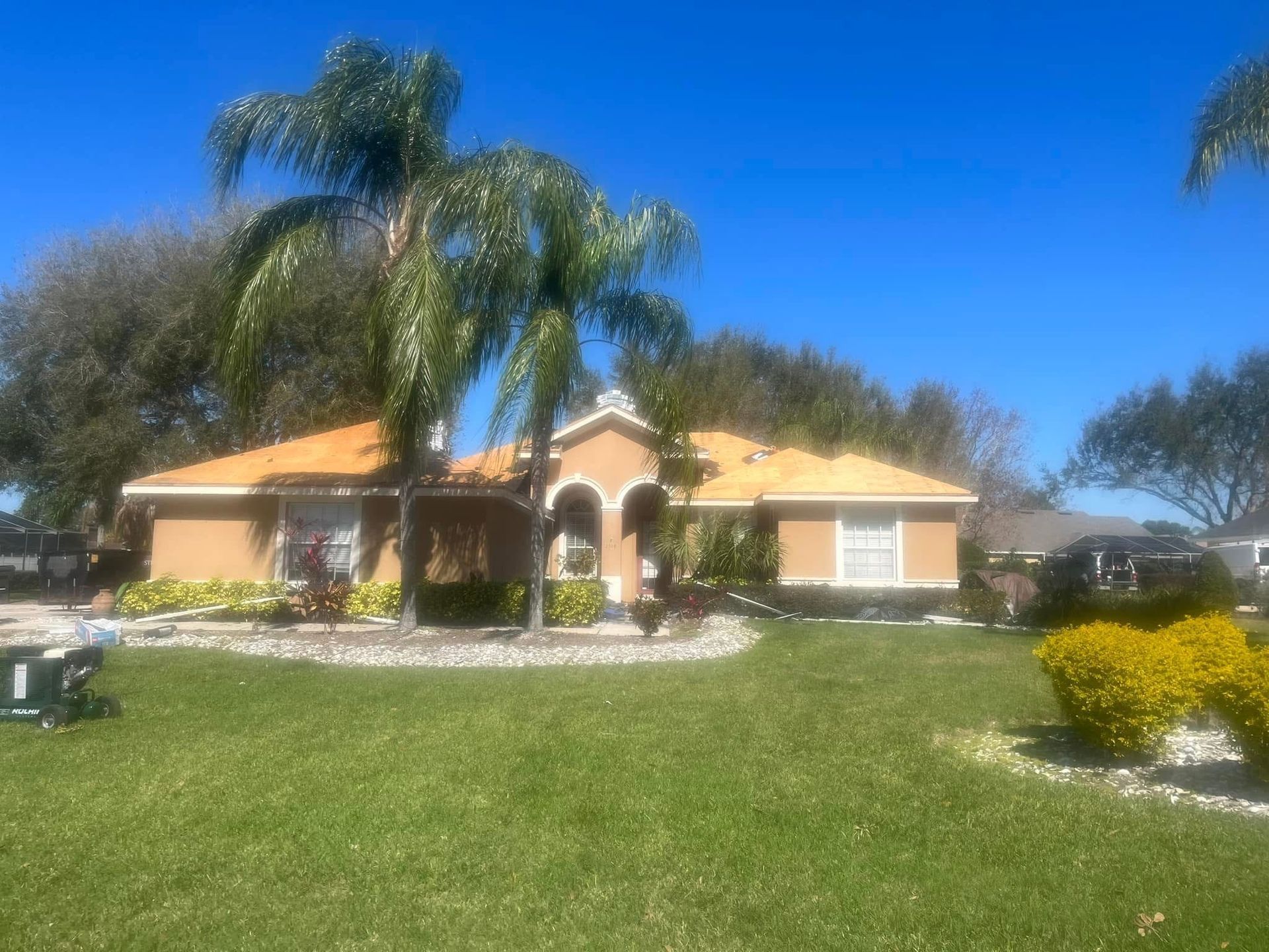 Tan house with orange roof, palm trees, and green lawn against a bright blue sky.