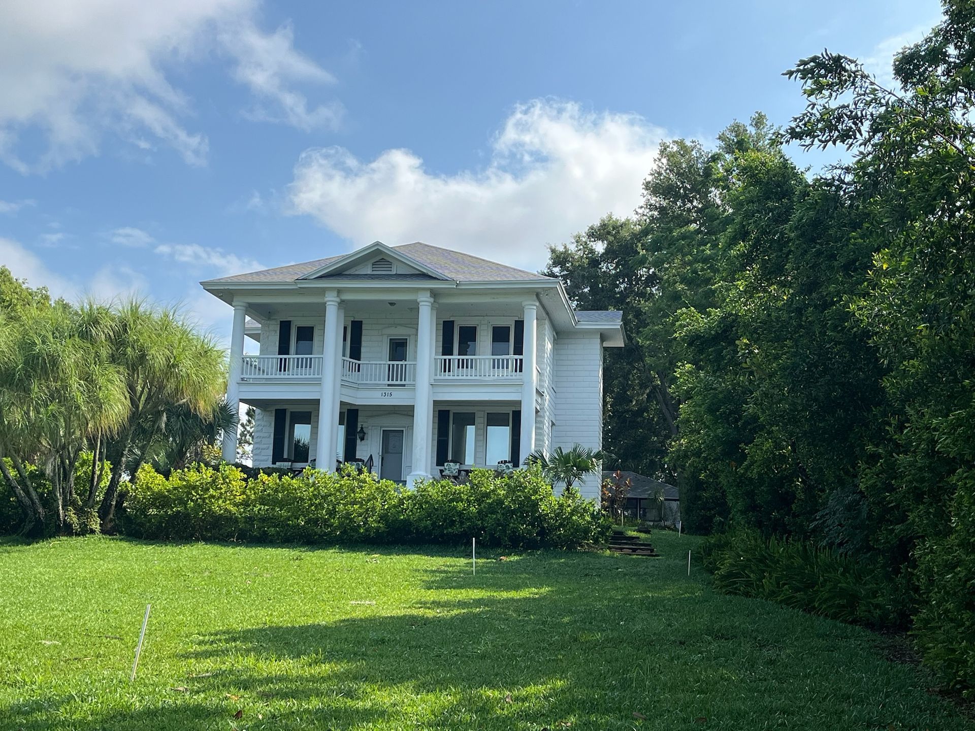White two-story house with columns and black shutters, set on a green lawn with trees, under a blue sky.