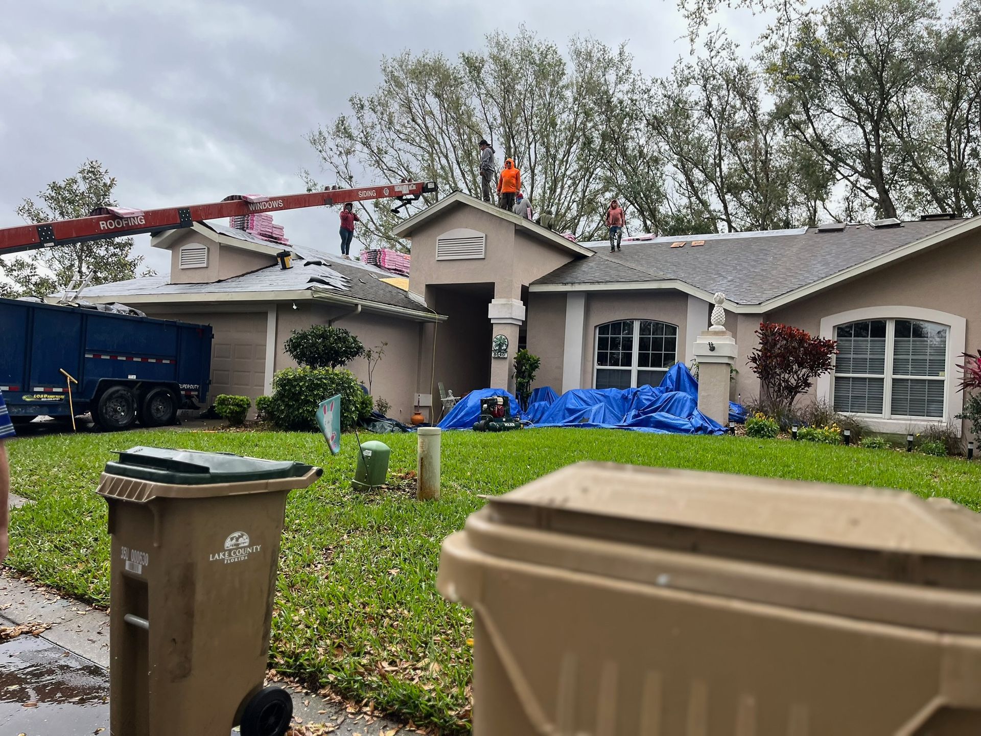 Roofers working on a house with a blue tarp, a dumpster truck, and two trash bins in the foreground.