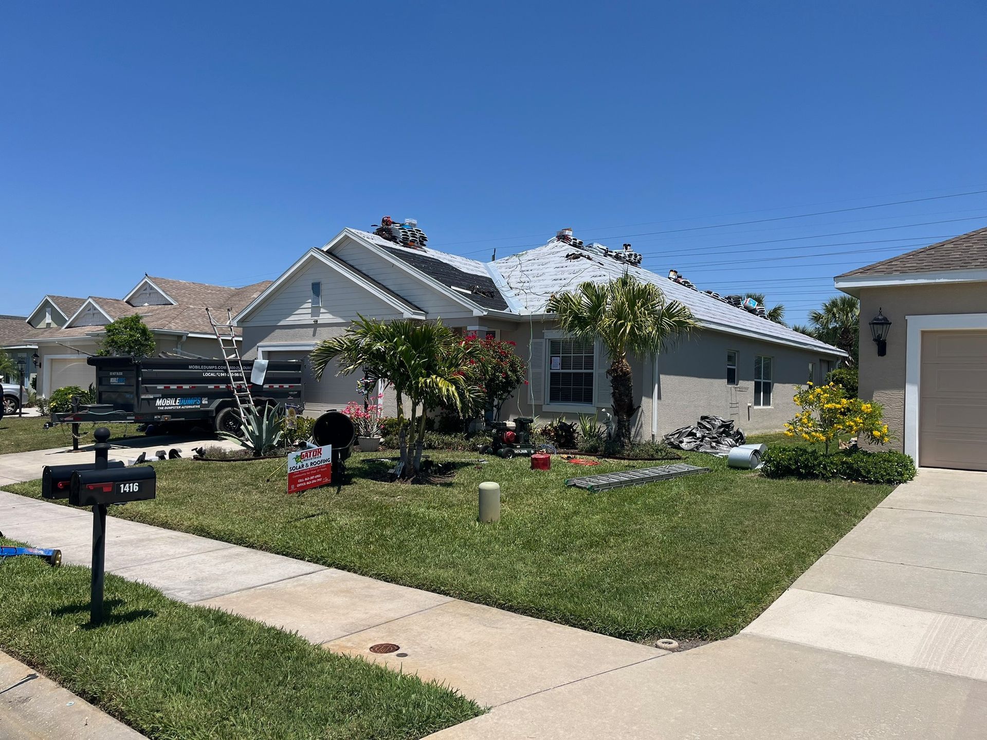 A house with workers on the roof under a clear blue sky; yard and surrounding houses are also visible.