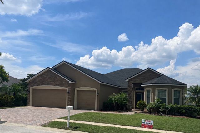 A tan, single-story house with a dark shingled roof, a paved driveway, and a small front lawn under a blue cloudy sky.