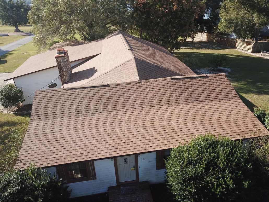 An aerial view of a single-story house with a brown shingled roof, a brick chimney, and surrounding trees and lawn.