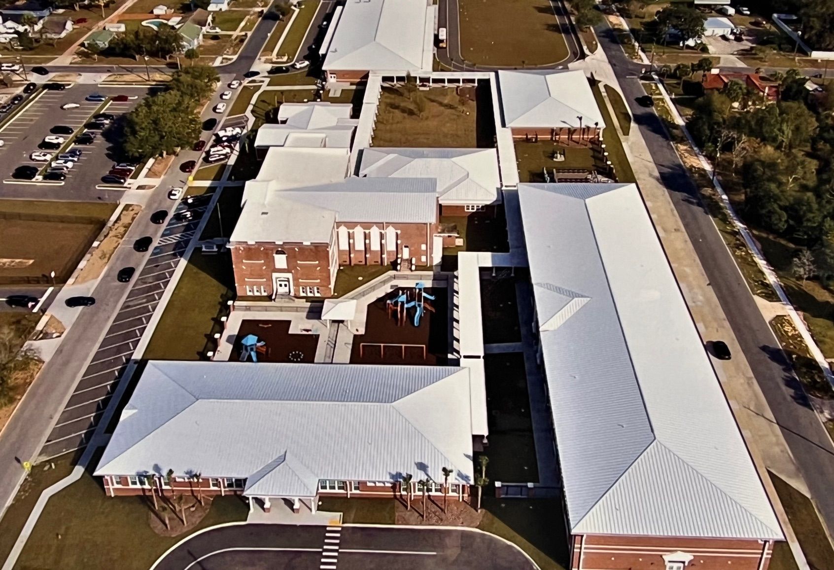 Aerial view of a school campus featuring brick buildings with light-colored metal roofs and an outdoor playground area.