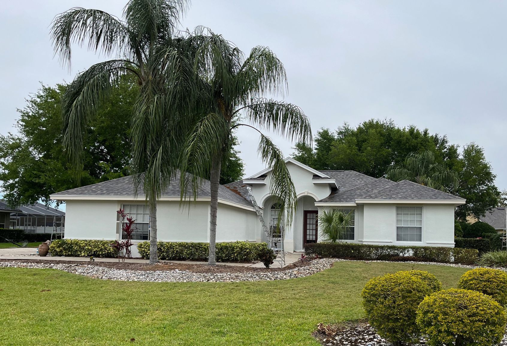 Single-story white suburban house with a tiled roof, front palm trees, and manicured landscaping under a cloudy sky.