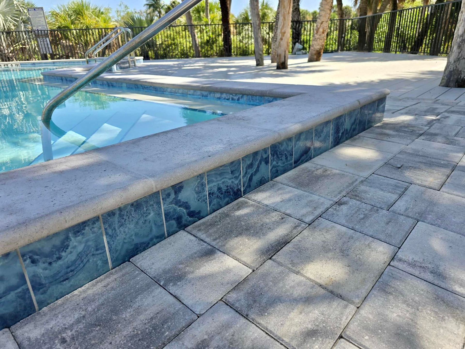 Swimming pool edge with blue tile, metal handrail, and stone-paved deck in sunlight