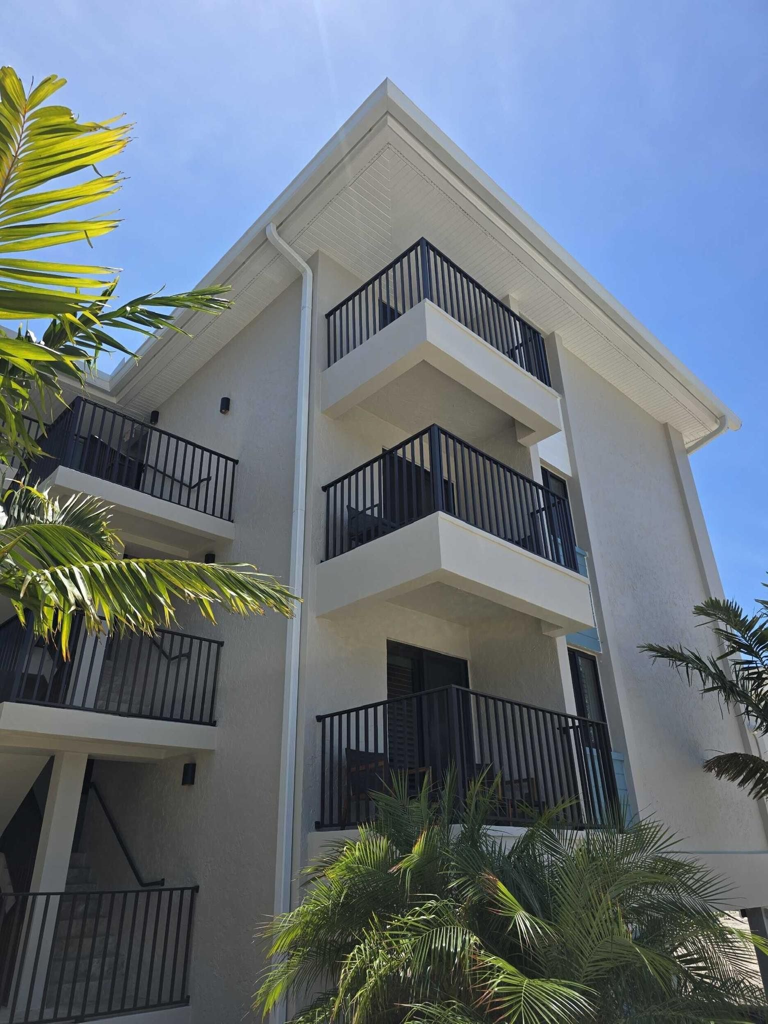 White apartment building with balconies and black railings, seen against a bright blue sky with palm trees.