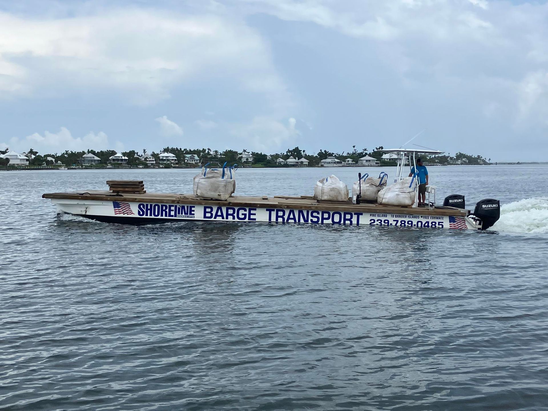 Long white transport barge on choppy water with bags and cargo, near a coastal village under cloudy sky