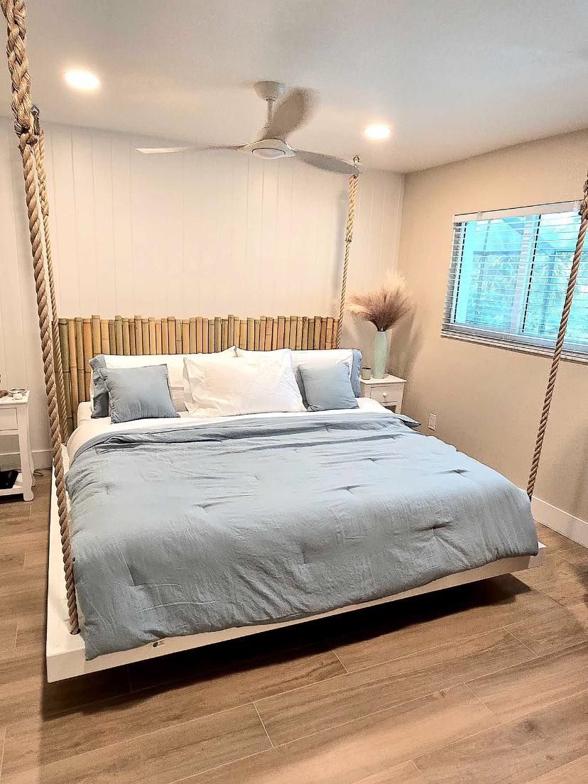 Neatly made bed with light blue bedding in a bright bedroom with a ceiling fan and window blinds