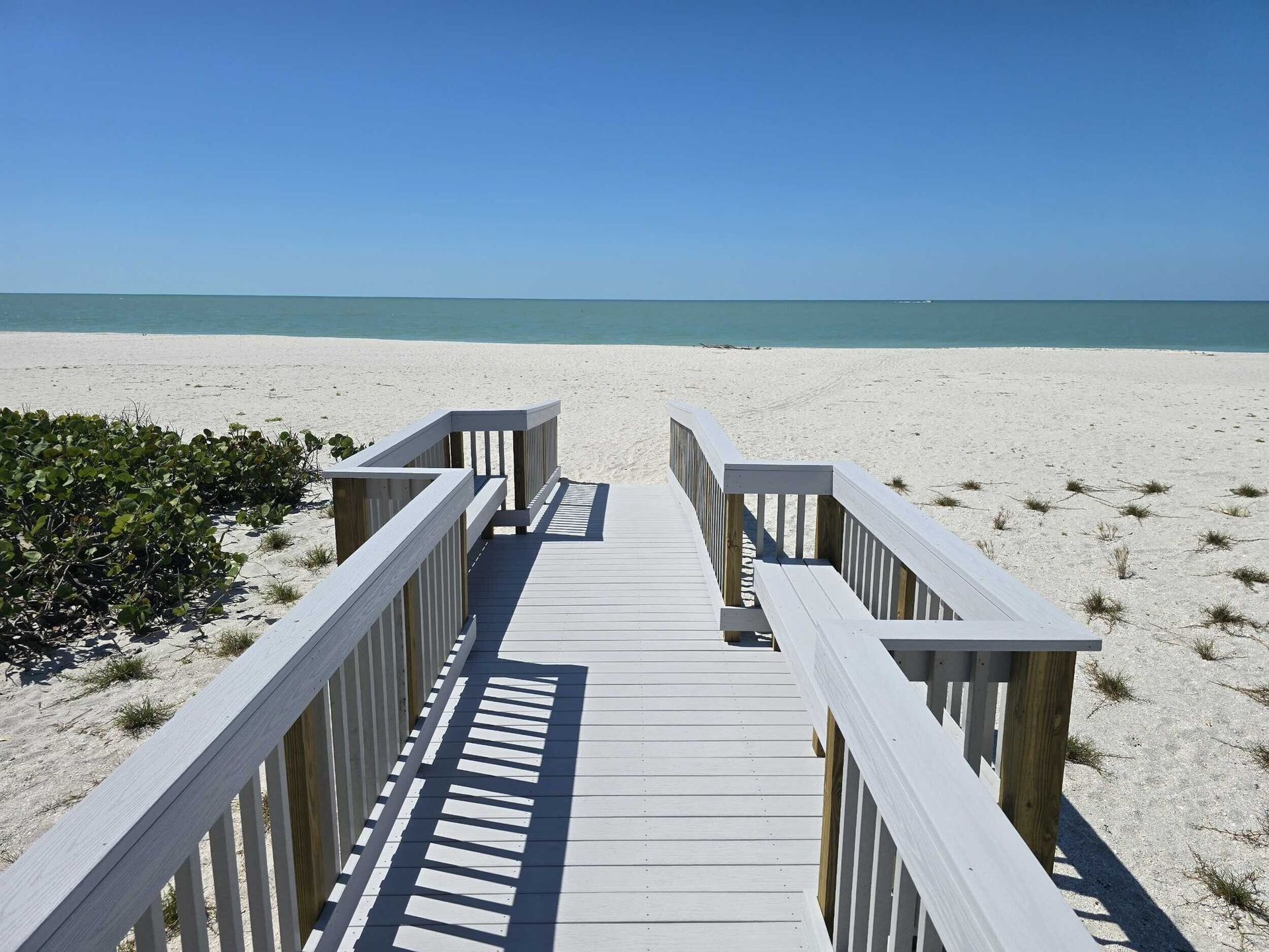 Wooden boardwalk leading to a sunny sandy beach and calm ocean under a clear blue sky