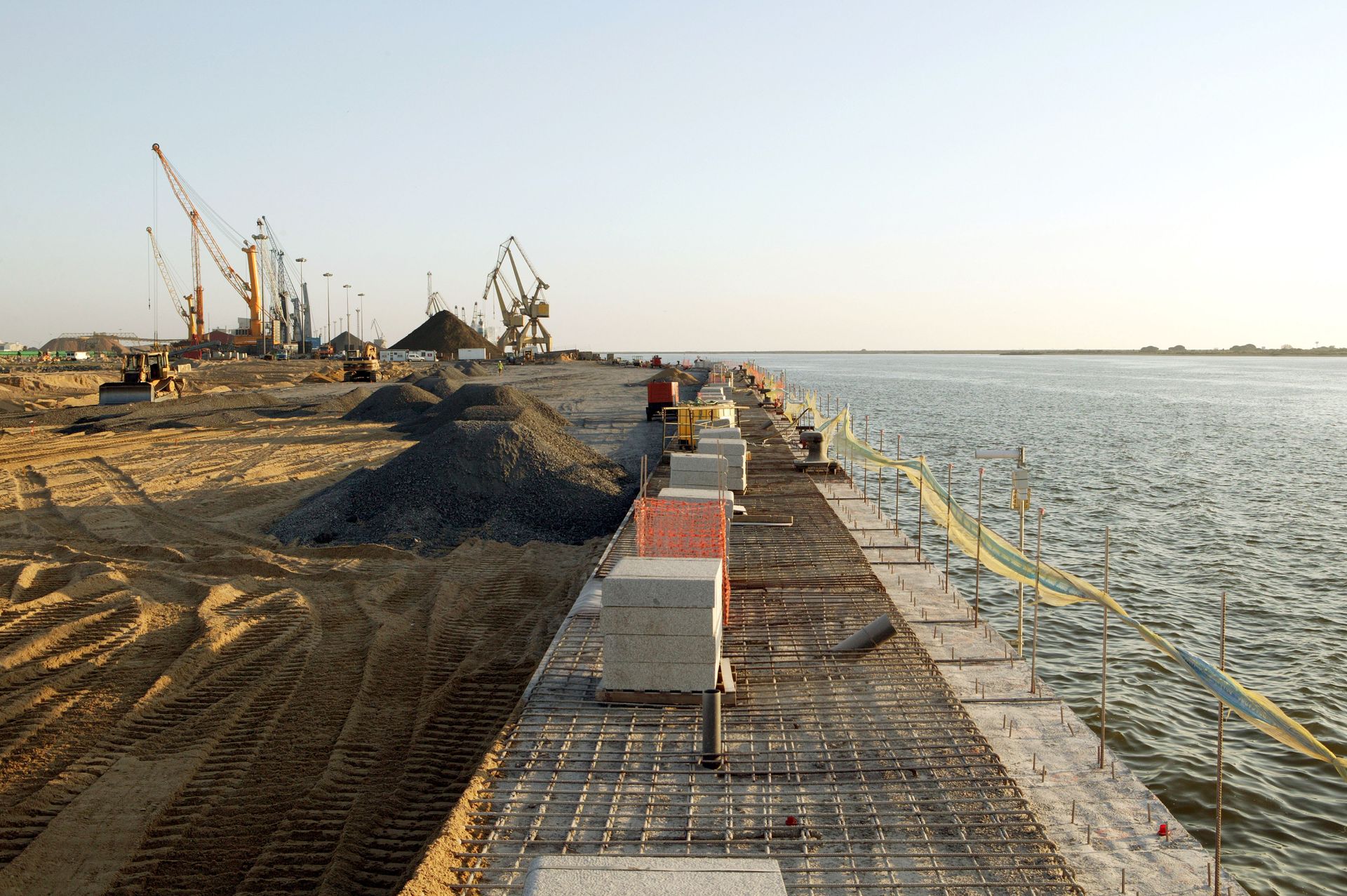 Construction site along a waterfront with cranes, barriers, and a paved path beside the water