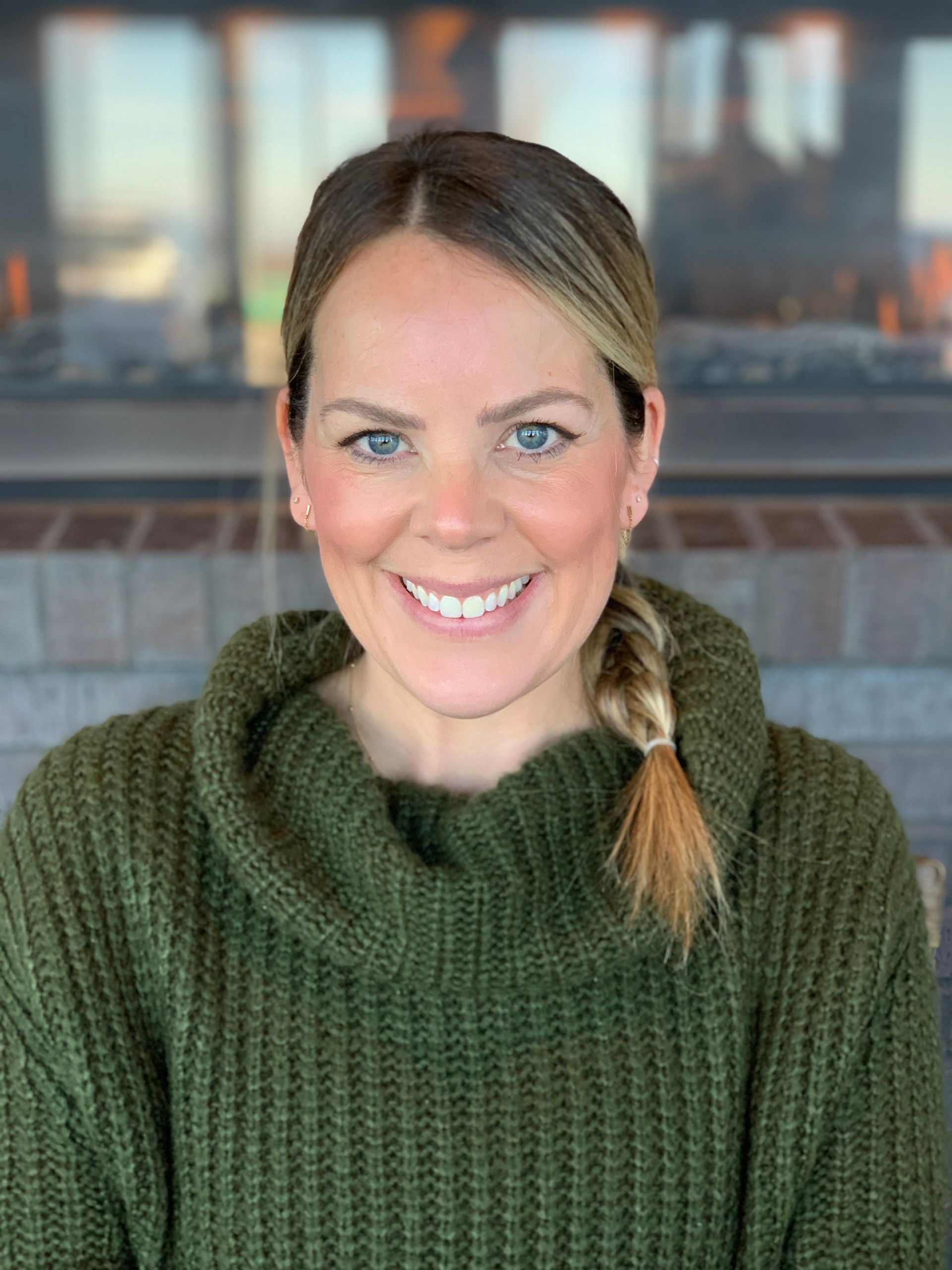 A smiling person with blonde-highlighted hair in a side braid, wearing a chunky olive green knit sweater by a fireplace.