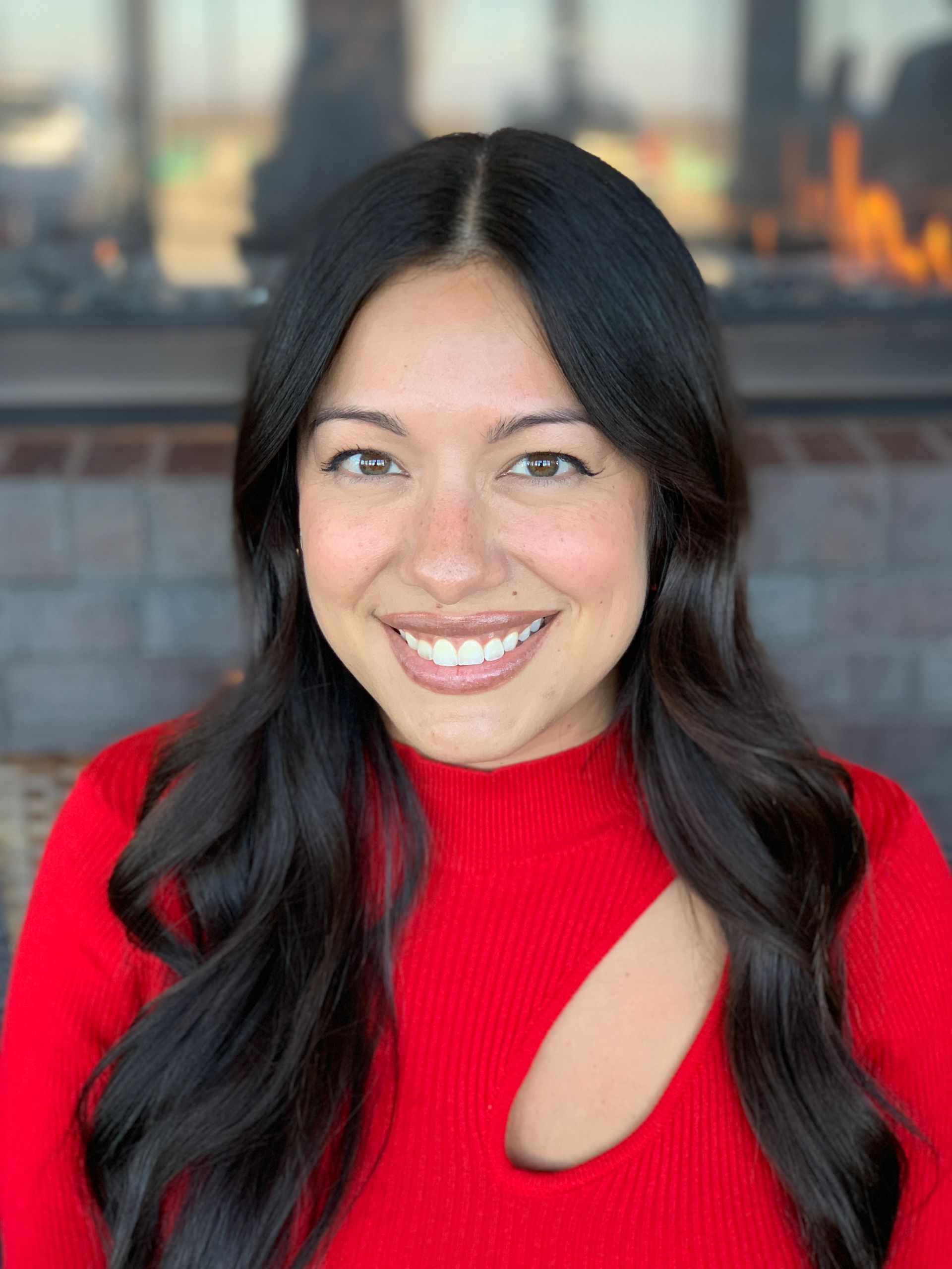 A person with long, dark hair wearing a bright red top with a cutout, smiling in front of an indoor fireplace.