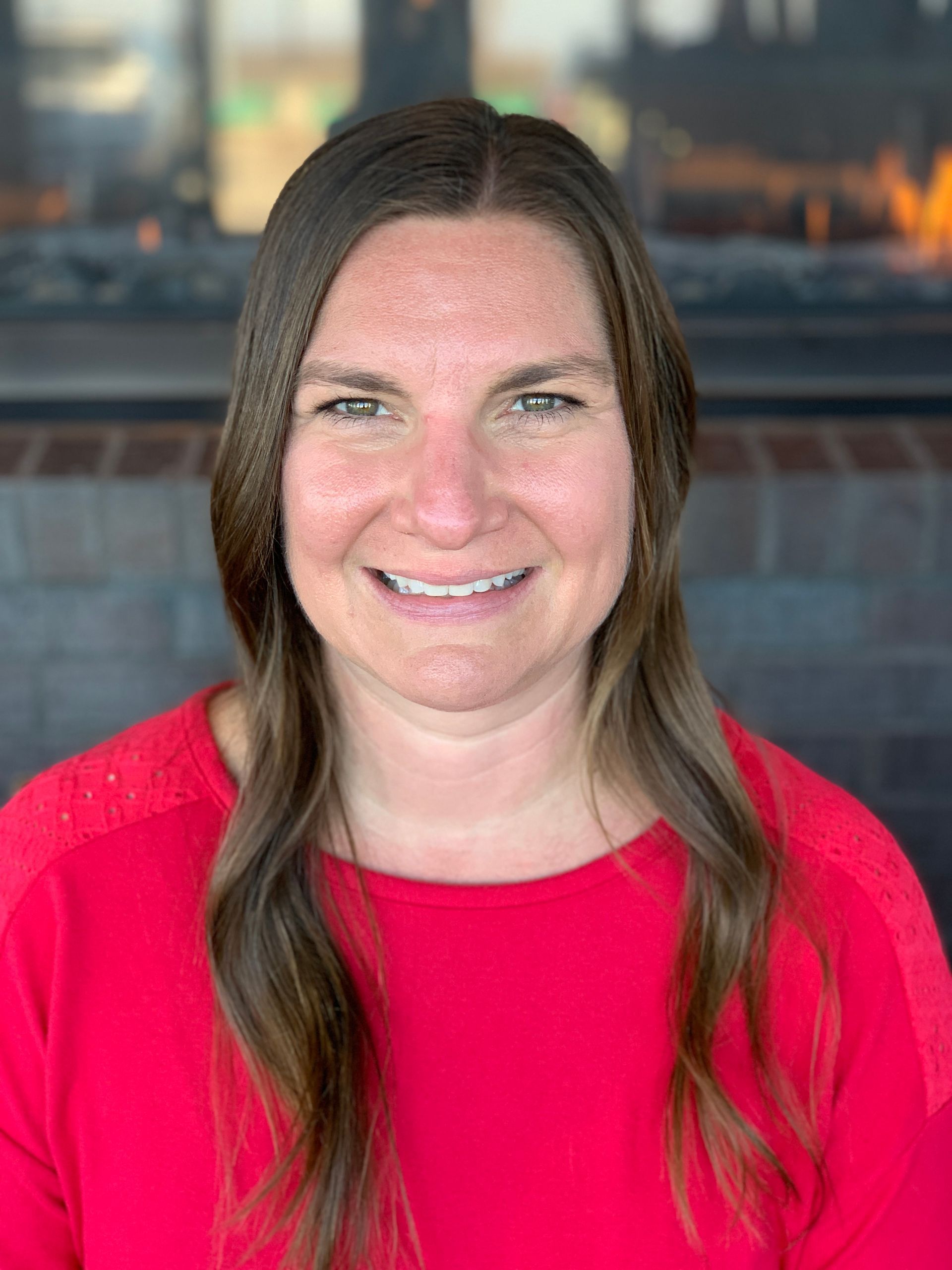 A person with long brown hair, smiling, wearing a red shirt in front of a brick fireplace.