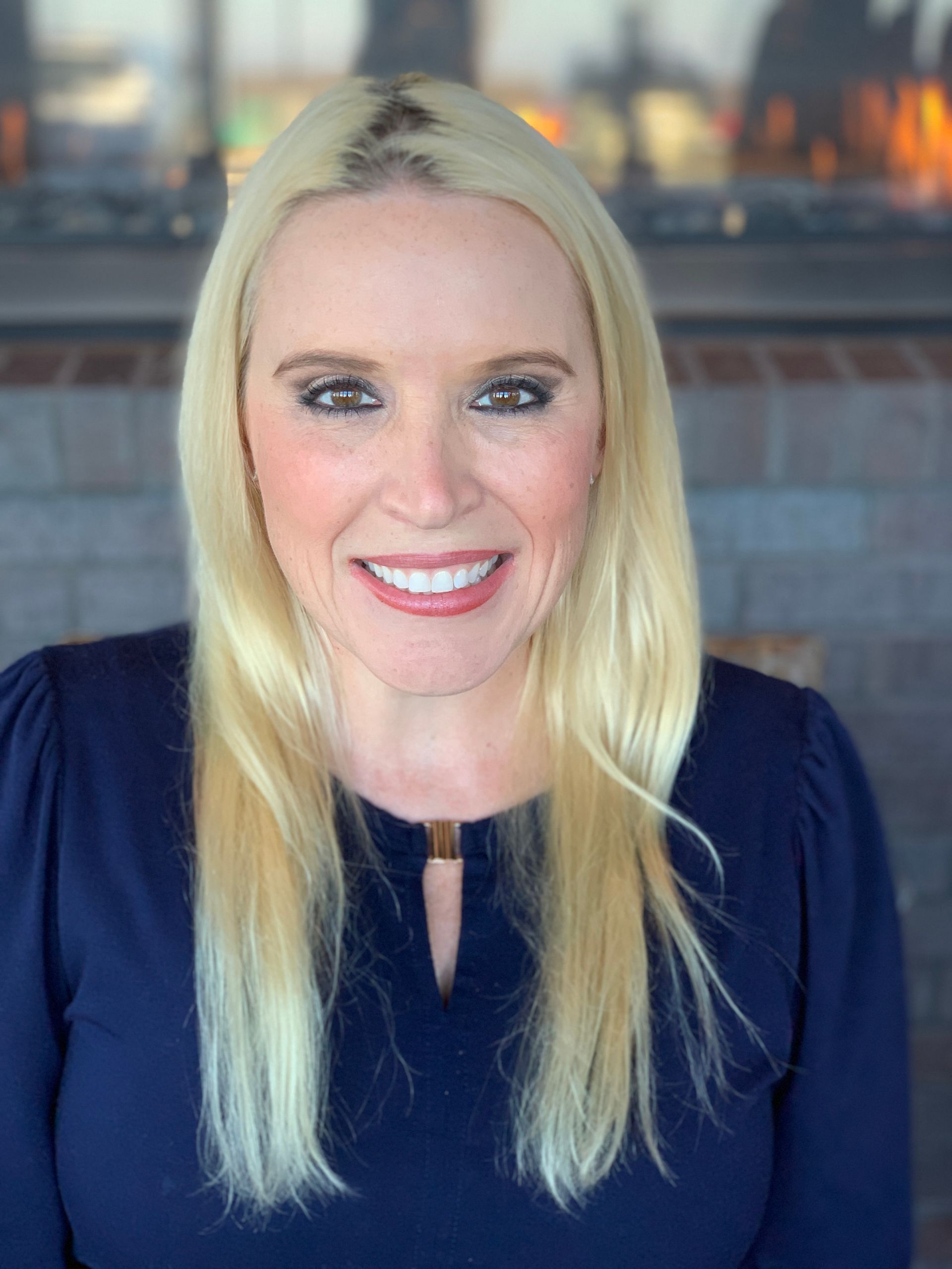 A smiling person with long blonde hair, wearing a navy blue long-sleeved top, posing in front of a blurred brick fireplace.