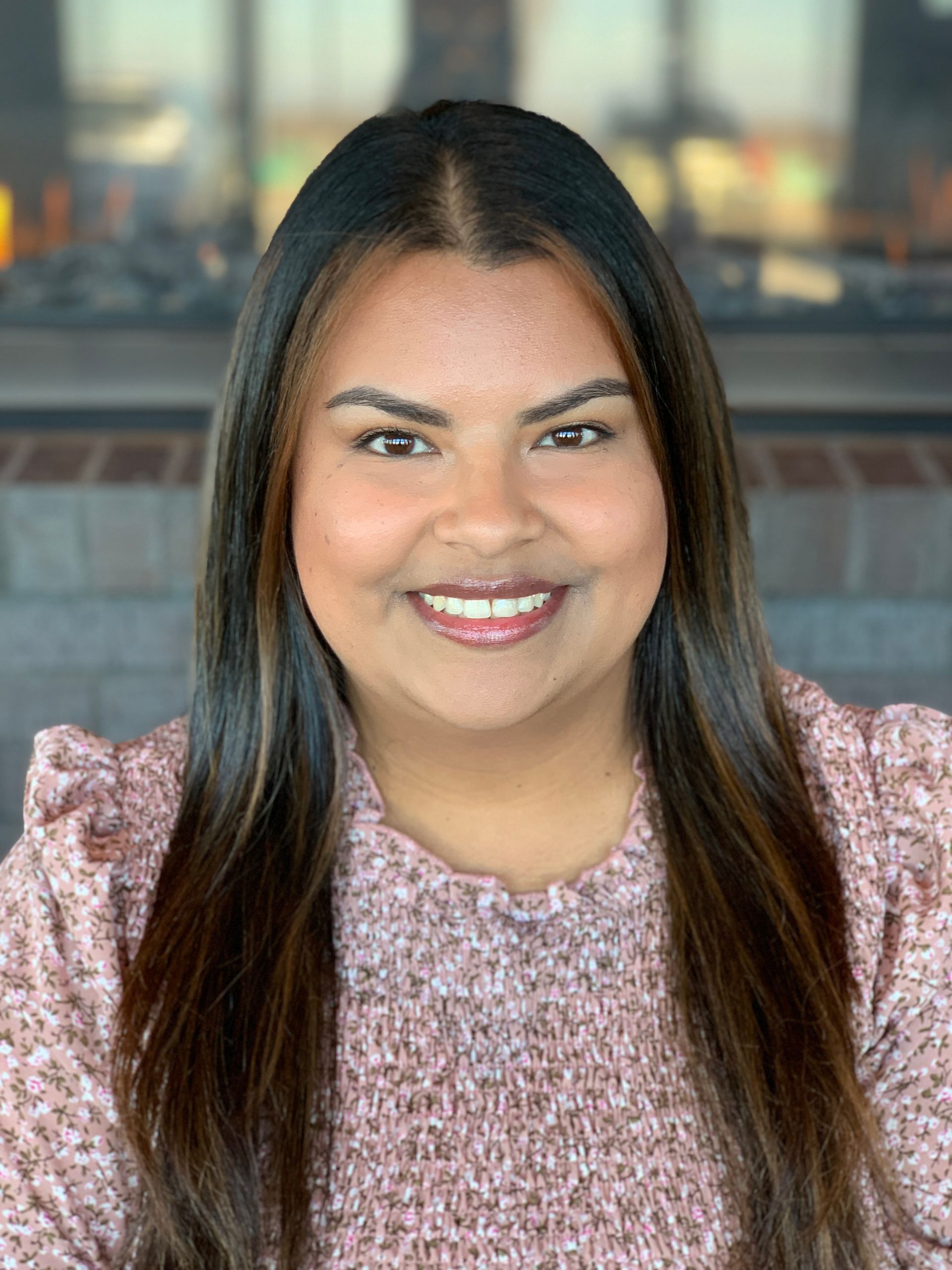 A smiling person with long dark hair wearing a light pink floral top, posed in front of a brick fireplace.