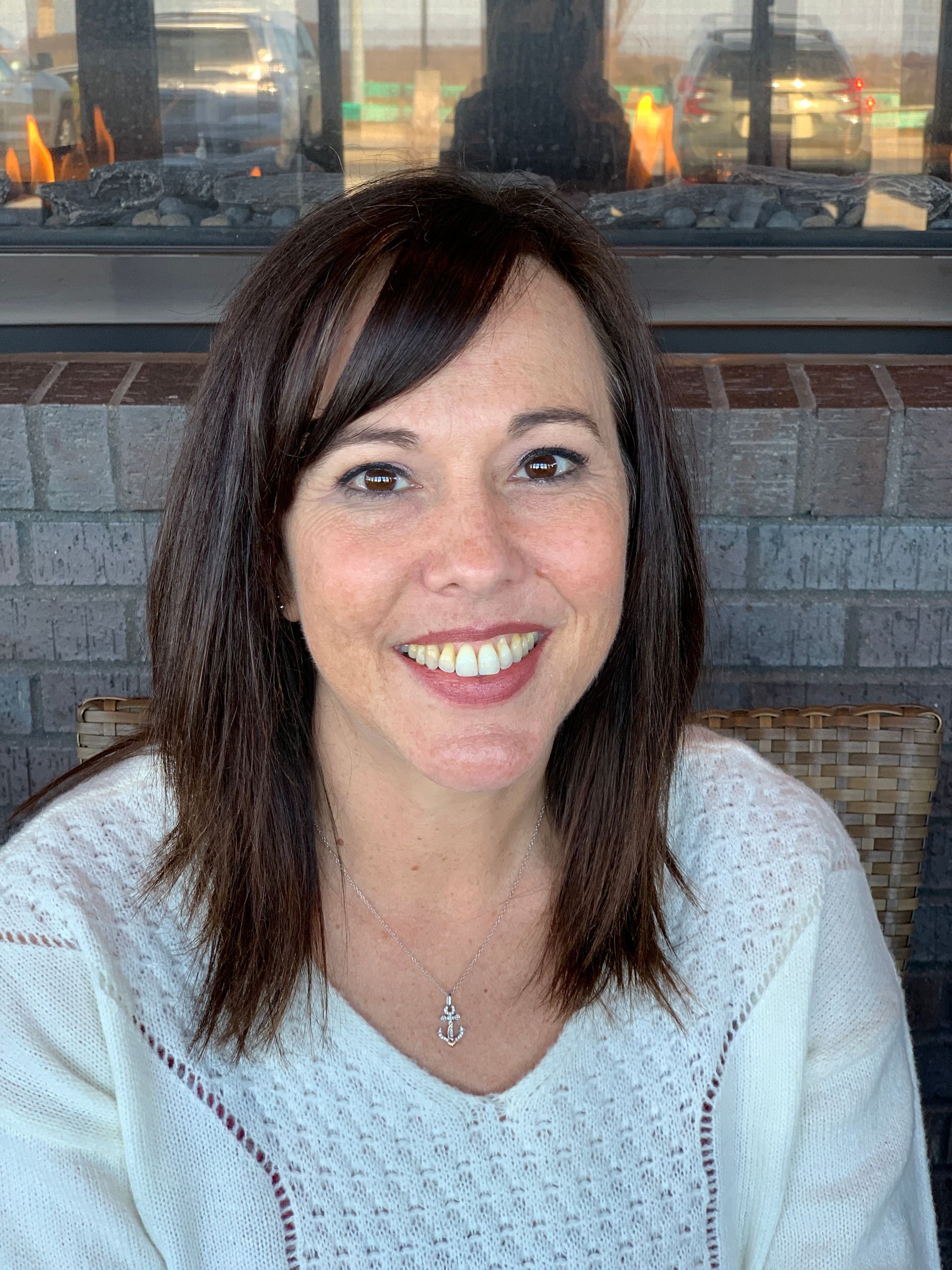 A smiling woman with shoulder-length dark hair wearing a white sweater, positioned in front of a brick fireplace.